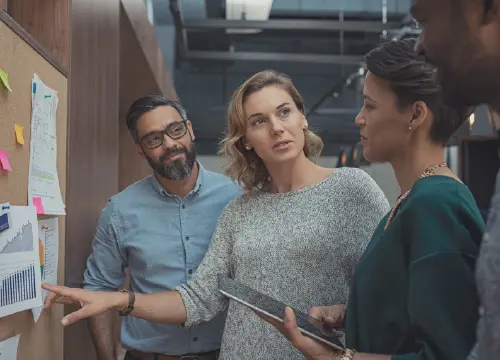Three workers, two women and one man, looking at information on a pinup board.