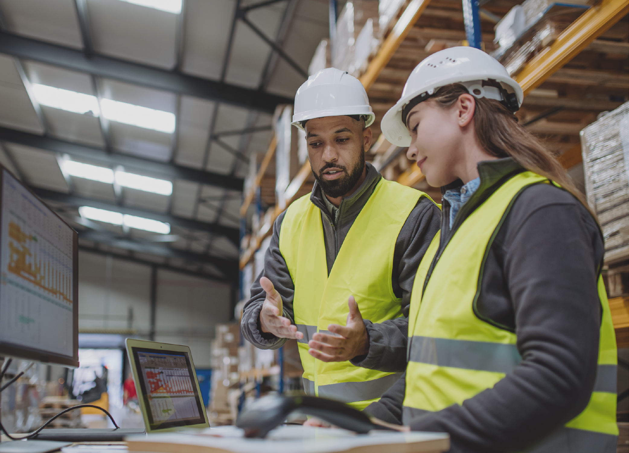 Man and a woman in hardhats and yellow vests working in a warehouse.