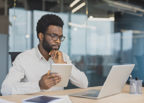 Male employee in glasses looking at his laptop screen while holding a tablet in his hand.