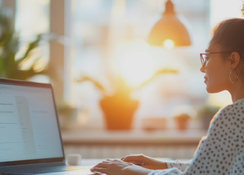 Woman working at a computer on a desk with sunlight coming through the window beside her.