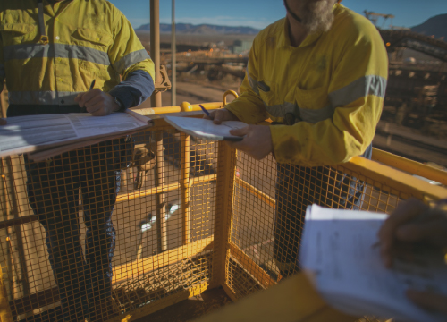 Two employees in yellow safety jackets up high collaborating.