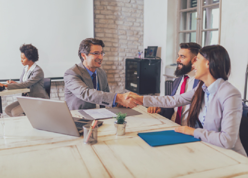 Three employees meeting around a table with two of them shaking hands.