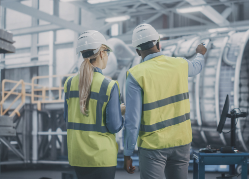 Two warehouse employees facing away wearing yellow safety vests and hard hats.