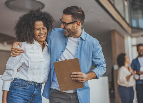 A male employees smiling with his arm around a smiling female employee while holding a clipboard.