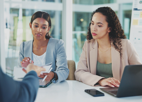 Two female employees at a table looking at something a third employee is handing them.