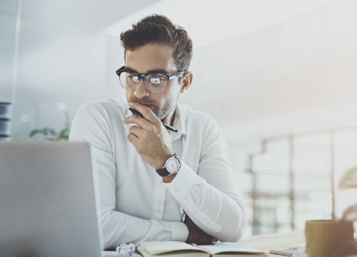 A man thinking about something while staring at a laptop screen with a notebook in front of him.