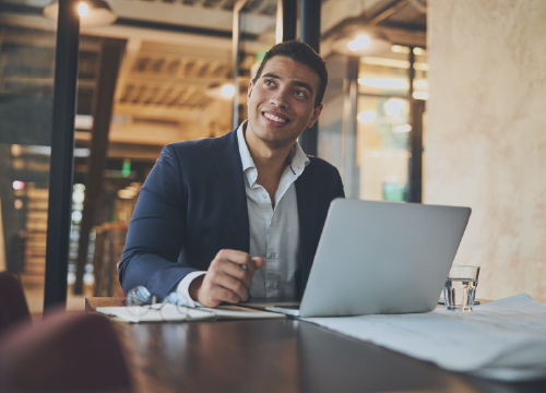 A man working at a table with his laptop and smiling at someone.