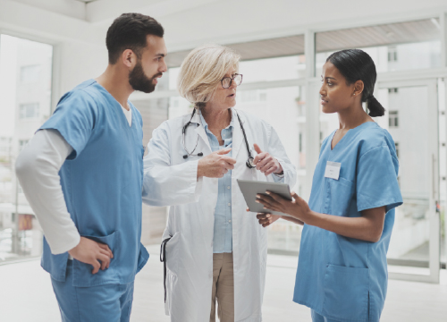 Three healthcare employees, two in blue scrubs and one in a lab coat talking.