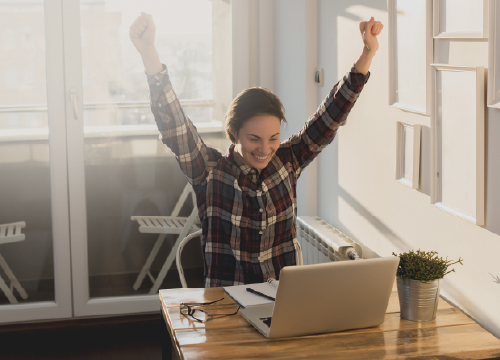 Female raising her arms in triumph sitting at a desk in front of a laptop.