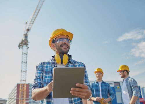 Three people working in a shipyard with a crane and supplies behind them.