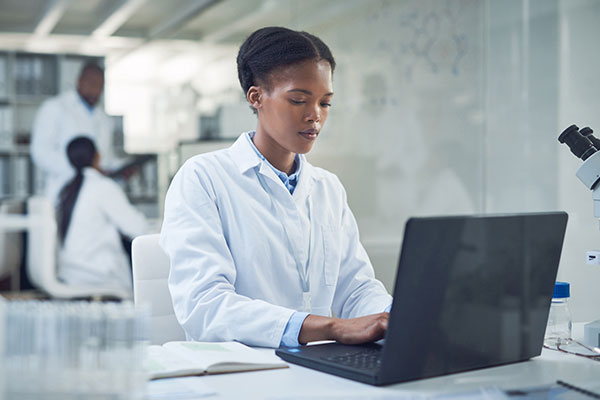 Female lab tech typing on a laptop wearing her lab coat.